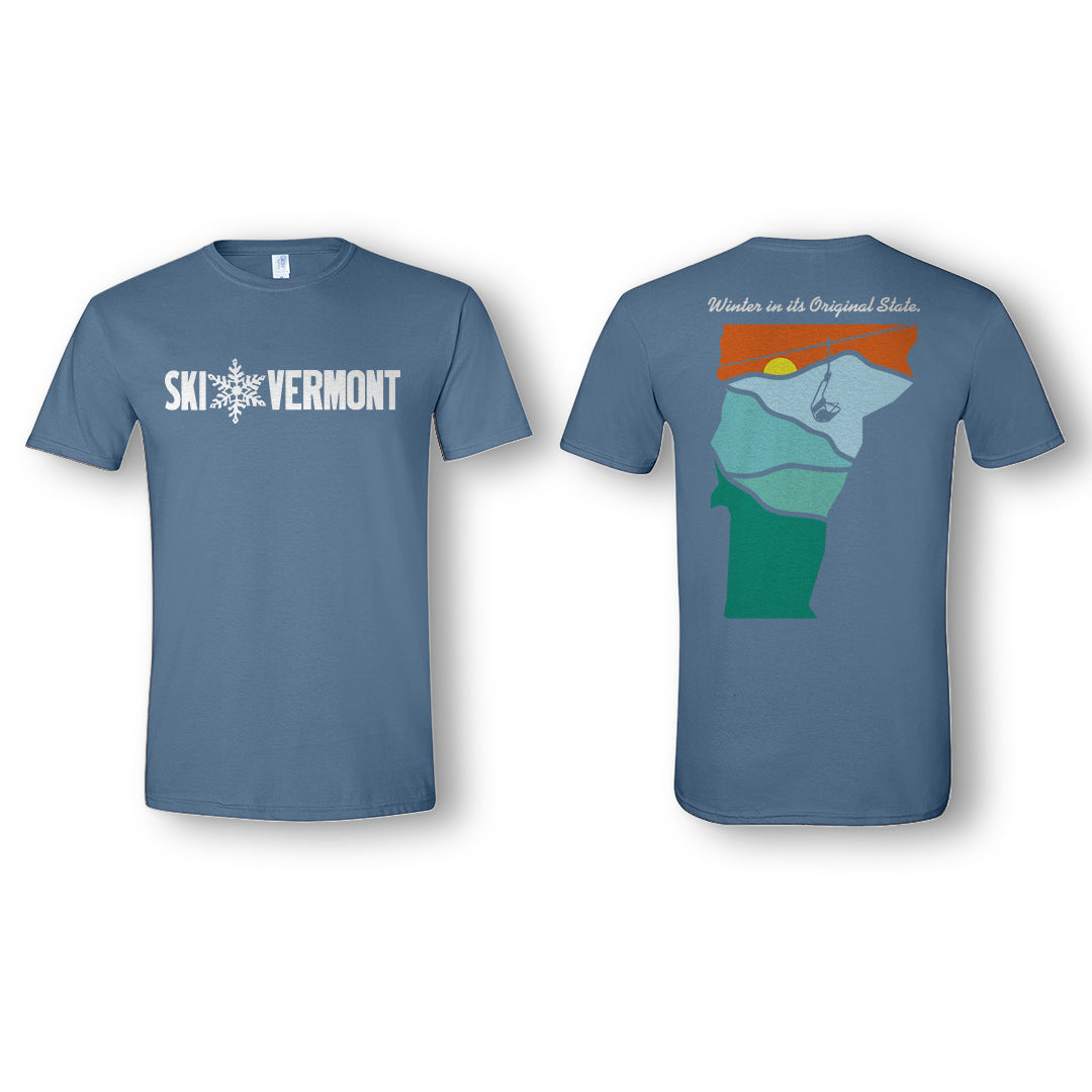 Front and back of a blue “Ski Vermont” T-shirt. The front features the Ski Vermont logo in white, with a snowflake design between the words “Ski” and “Vermont.” The back shows a stylized outline of the state of Vermont filled with layered landscape colors — green, blue, and orange — depicting mountains, a sunset, and a ski lift. Above the design, white text reads “Winter in its Original State.”